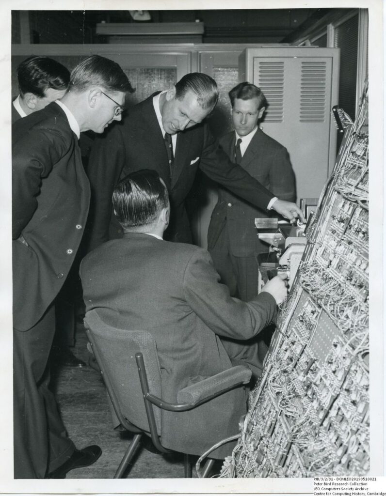 Prince Philip asking a question about the operation of the LEO III wiring plane and paper tape reader. Standing to the left is John Pinkerton. Taken at LEO's computer works and factory on Minerva Road in Acton, London 22 March 1960